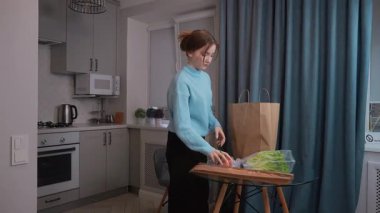 Genç bir kız, şık ve modern bir mutfakta, kese kağıdından taze ürünler ve diğer yiyecek eşyaları çıkarıyor, sağlıklı yaşam tarzını ve ev hayatını ön plana çıkarıyor.