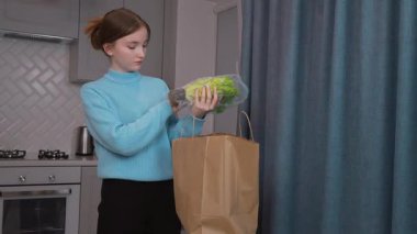 Genç kız, kese kağıdından paketleri boşalttıktan sonra mutfak masasına taze yiyecekler seriyor, yeniden kullanılabilir ağ torbalarının etrafını organik meyve ve sebzelerle çeviriyor. dikey video