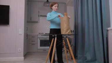 Genç kız, kese kağıdından paketleri boşalttıktan sonra mutfak masasına taze yiyecekler seriyor, yeniden kullanılabilir ağ torbalarının etrafını organik meyve ve sebzelerle donatıyor..