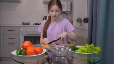 Yetenekli genç kız, modern mutfak ortamında ev yapımı salata için taze sebze düzenlerken ahşap kesme tahtasının üzerinde kırmızı soğan dilimliyor.