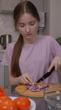 Genç kız ahşap kesim tahtasında kırmızı soğan doğruyor, modern mutfağında marul ve dereotla taze ve sağlıklı salata hazırlıyor.