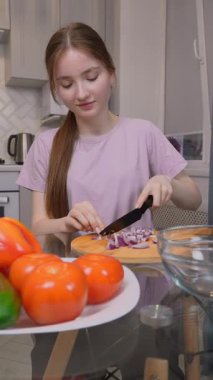 Genç kız ahşap kesim tahtasında kırmızı soğan doğruyor, modern mutfağında marul ve dereotla taze ve sağlıklı salata hazırlıyor.