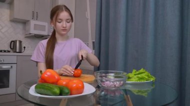 Genç kız domatesleri ahşap bir kesim tahtasında doğruyor. Etrafı marul, salatalık, dolma biber ve soğan gibi taze sebzelerle çevrili. Ev mutfağında sağlıklı bir salata hazırlıyorlar.