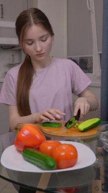 Genç kız, domates, biber ve marul gibi taze sebzelerle çevrili ahşap kesim tahtasının üzerinde salatalık doğruyor.