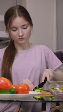 Genç bir kız cam masada salatalık doğruyor, taze, besleyici bir salata hazırlıyor, renkli sebzelerle, parlak ev mutfağında dikey video kasette.