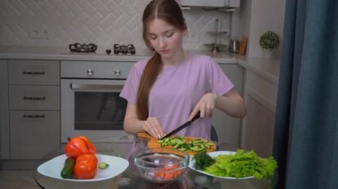 Genç bir kız cam masada salatalık doğruyor, taze, besleyici bir salata hazırlıyor, renkli sebzelerle, parlak mutfak işyerinde.