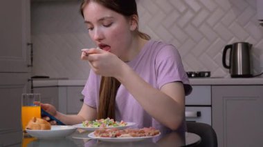 Genç bir kız kahvaltı yaparken cam masada oturuyor ve sakin bir sabah ortamında portakal suyu, ekmek ve salatanın tadını çıkarıyor.