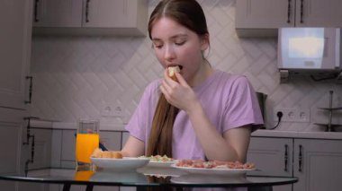 Genç kız modern mutfakta jambonlu sandviç yapıyor, masada oturuyor, taze sıkılmış portakal suyu içiyor ve sabah kahvaltısının tadını çıkarıyor.