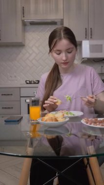 Sevimli bir genç kız kahvaltıda meyve suyu yer ve akıllı telefon ekranı seyreder. Mutfak masasında kameranın önünde oturur ve çevrimiçi video izler.