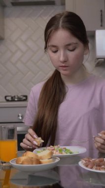 Sevimli bir genç kız kahvaltıda meyve suyu yer ve akıllı telefon ekranı seyreder. Mutfak masasında kameranın önünde oturur ve çevrimiçi video izler.