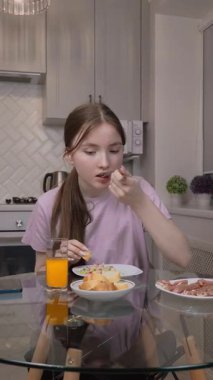 Sevimli bir genç kız kahvaltıda meyve suyu yer ve akıllı telefon ekranı seyreder. Mutfak masasında kameranın önünde oturur ve çevrimiçi video izler.