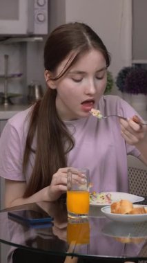 Genç kız evde besleyici yiyeceklerin tadını çıkarırken gündelik bir öğle yemeği videosunda modern bir mutfakta oturmuş salata, jambon, ekmek ve portakal suyu yiyor.