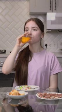 Genç kız portakal suyu içiyor ve modern mutfakta salata, jambon ve ekmekten sağlıklı bir kahvaltı yiyor. Dengeli bir yaşam tarzı ve sabah videosu gösteriyor.