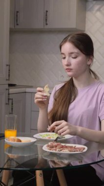 Uzun kahverengi saçlı, günlük mor tişörtlü genç kız modern mutfakta cam masada oturmuş kurutulmuş et, salata, ekmek ve portakal suyu dikey videosunun tadını çıkarıyor.