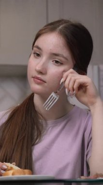 Genç bir kız mutfak masasında elinde çatalla oturuyor ve düşüncelerinde kaybolmuş gibi görünüyor. Belki de kahvaltıda endişelerle ya da üzüntüyle uğraşıyordur.