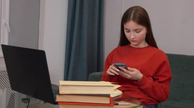 Kırmızı kazaklı, motive olmuş genç bir öğrenci, yoğun bir şekilde çalışıyor, akıllı telefondaki dijital içerikte geziniyor ve ders kitapları ve dizüstü bilgisayarlarla çevrili olarak ev masasında çalışıyor.