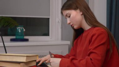 Genç kız masada oturuyor, dizüstü bilgisayar kullanıyor ve not alıyor, ergonominin önemini vurguluyor. Sınavlar ya da evde eğitim hazırlıkları.