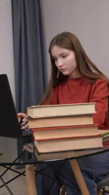 Cam masada özenle dizüstü bilgisayarda yazan odaklanmış kız öğrenci, kitap yığınının yanında odaklanmış çalışma ve akademik uğraşları gösteren bir yığın var. dikey video