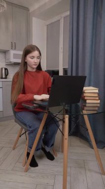 Genç kız öğrenci ders çalışmaya odaklanmış, dizüstü bilgisayar ve ders kitabı kullanıyor cam mutfak masasında oturuyor, günlük kırmızı kazak ve mavi kot dikey video kotlar giyiyor.