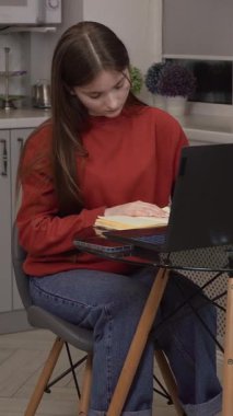 Genç kız öğrenci ders çalışmaya odaklanmış, dizüstü bilgisayar ve ders kitabı kullanıyor cam mutfak masasında oturuyor, günlük kırmızı kazak ve mavi kot dikey video kotlar giyiyor.