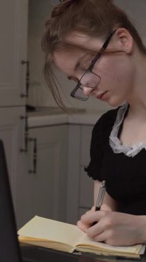 Bilgisayarla çevrimiçi çalışırken not alan odaklanmış öğrenci, evde otururken doğal ışık ve minimalist iç mekan dikey video ile bilgisayarını kullanıyor.