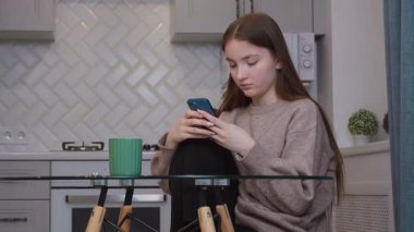 Genç kız, cam mutfak masasında yeşil seramik bardağın yanında otururken, akıllı telefonları hafifçe kaydırıyor.