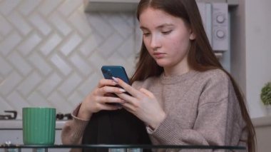 Genç kız, cam mutfak masasında yeşil seramik bardağın yanında otururken, akıllı telefonları hafifçe kaydırıyor.