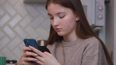 Genç kız, cam mutfak masasında yeşil seramik bardağın yanında otururken, akıllı telefonları hafifçe kaydırıyor.