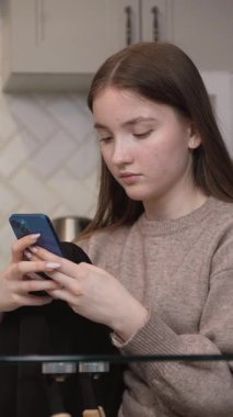 Konsantre genç kız internete veya sosyal medyaya girip cep telefonunu kullanarak ev mutfağındaki cam masada oturuyor ve dikey giysiler giyiyor.