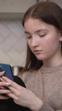 Konsantre genç kız internete veya sosyal medyaya girip cep telefonunu kullanarak ev mutfağındaki cam masada oturuyor ve dikey giysiler giyiyor.