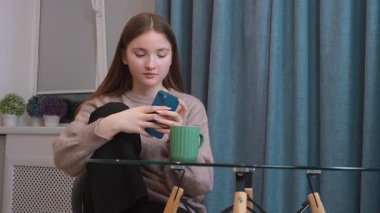 Genç kız, cam mutfak masasında yeşil seramik bardağın yanında otururken, akıllı telefonları hafifçe kaydırıyor.