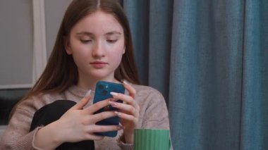 Genç kız dinleniyor, akıllı telefonla sohbet ediyor, sosyal medya uygulamalarına kayıyor, arkadaşlarına mesaj atıyor, rahat bir ev ortamında çevrimiçi video izliyor.