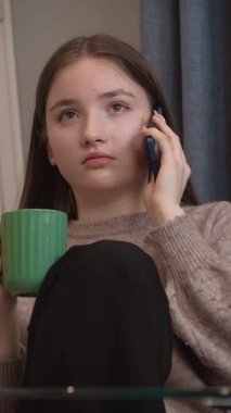 Genç kız, rahat bir şekilde mutfakta oturuyor, yeşil bardaktan çay içiyor ve telefon görüşmesi yapıyor, yüz ifadelerinin dinamik aralığını ve dikey iletişim sinyallerini gösteriyor.