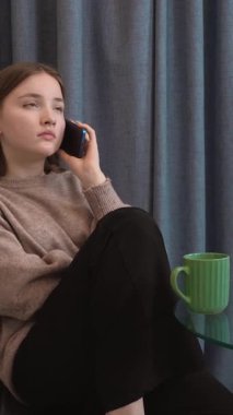 Genç kız, rahat bir şekilde mutfakta oturuyor, yeşil bardaktan çay içiyor ve telefon görüşmesi yapıyor, yüz ifadelerinin dinamik aralığını ve dikey iletişim sinyallerini gösteriyor.