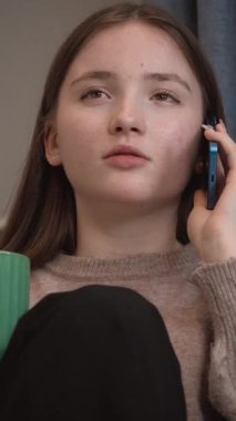 Genç kız, rahat bir şekilde mutfakta oturuyor, yeşil bardaktan çay içiyor ve telefon görüşmesi yapıyor, yüz ifadelerinin dinamik aralığını ve dikey iletişim sinyallerini gösteriyor.