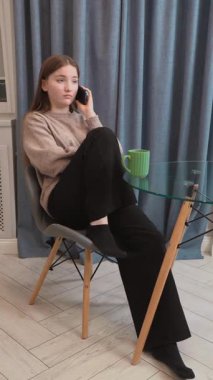 Genç kız cam masanın yanında dinleniyor, rahat bir telefon görüşmesi yapıyor, elinde yeşil bir kupa tutuyor ve sakin bir ev ortamını yumuşak, rahat bir atmosfer dikey video ile somutlaştırıyor.