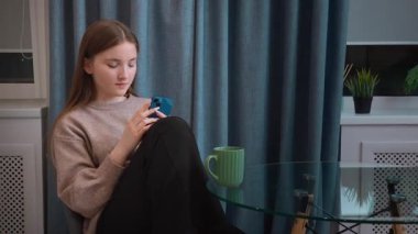 Genç kız cam masanın yanında dinleniyor, rahat bir telefon görüşmesi yapıyor, elinde yeşil kupa tutuyor ve sakin bir ev ortamını yumuşak, rahat bir atmosferle somutlaştırıyor.