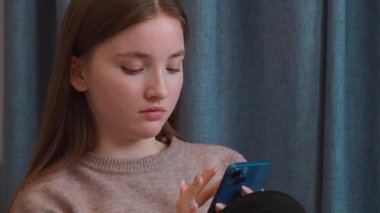 Oturma odasında oturan genç kız akıllı telefon ekranını izlerken karışık duygular yaşıyor. Yüzünde, elinde yeşil kahve fincanıyla düşünceli ve şaşırmış bir ifade var.