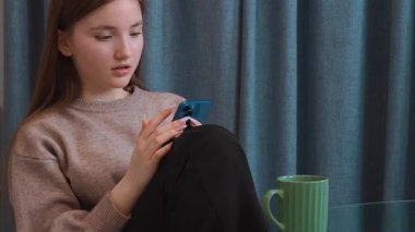 Oturma odasında oturan genç kız akıllı telefon ekranını izlerken karışık duygular yaşıyor. Yüzünde, elinde yeşil kahve fincanıyla düşünceli ve şaşırmış bir ifade var.