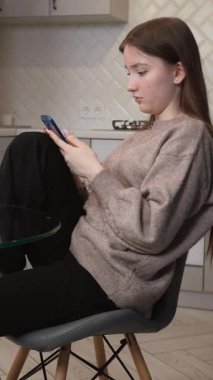 Oturma odasında oturan genç kız akıllı telefon ekranını izlerken karışık duygular yaşıyor. Yüzünde, elinde yeşil kahve fincanıyla düşünceli ve şaşırmış bir ifade var.