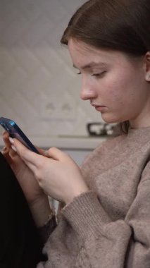 Oturma odasında oturan genç kız akıllı telefon ekranını izlerken karışık duygular yaşıyor. Yüzünde, elinde yeşil kahve fincanıyla düşünceli ve şaşırmış bir ifade var.