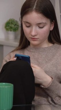 Genç bir kız mavi akıllı telefonuyla sosyal medyada gezinirken cam bir masada yeşil kurdeleli camların yanında, konsantre yüz ifadesi olan dijital içeriğe odaklanıyor.