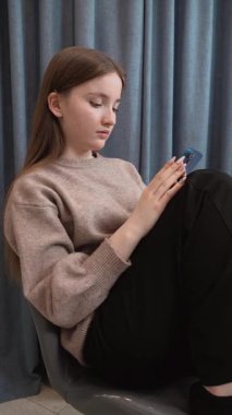 Genç bir kız mavi akıllı telefonuyla sosyal medyada gezinirken cam bir masada yeşil kurdeleli camların yanında, konsantre yüz ifadesi olan dijital içeriğe odaklanıyor.