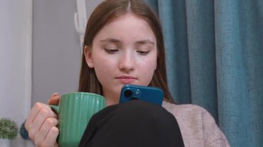 Genç bir kadın evinde dinleniyor, akıllı telefona bakarken sıcak bir içecek yudumluyor, dijital çağın rahatlığını ve modern yaşam tarzının bağlantısını somutlaştırıyor.