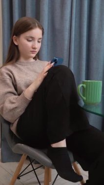 Genç bir kız mavi akıllı telefonuyla sosyal medyada gezinirken cam bir masada yeşil kurdeleli camların yanında, konsantre yüz ifadesi olan dijital içeriğe odaklanıyor.