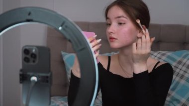 Profesyonel güzellik etkeni genç dikkatlice makyaj uyguluyor. Yüzük ışığı, akıllı telefon ve küçük ayna kullanarak göz alıcı çevrimiçi içerik yaratıyor.