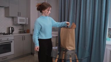 Genç kız kese kağıdından taze yiyecekler çıkarıyor, marul, meyve suyu ve sütü düzgünce modern mutfak eko torbalarında cam masaya yerleştiriyor.