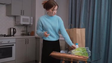 Genç kız kese kağıdından taze yiyecekler çıkarıyor, marul, meyve suyu ve sütü düzgünce modern mutfak eko torbalarında cam masaya yerleştiriyor.