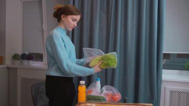 Günlük kıyafetli genç bir kız market alışveriş listesini kontrol ediyor. Mutfak masasındaki çevre dostu yeniden kullanılabilir çantalardan taze ürünler ve paketlenmiş gıdalar çıkarıyor.