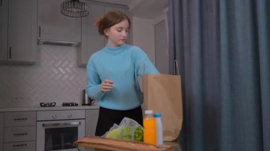 Genç kız kese kağıdından taze yiyecekler çıkarıyor, marul, meyve suyu ve sütü düzgünce modern mutfak eko torbalarında cam masaya yerleştiriyor.